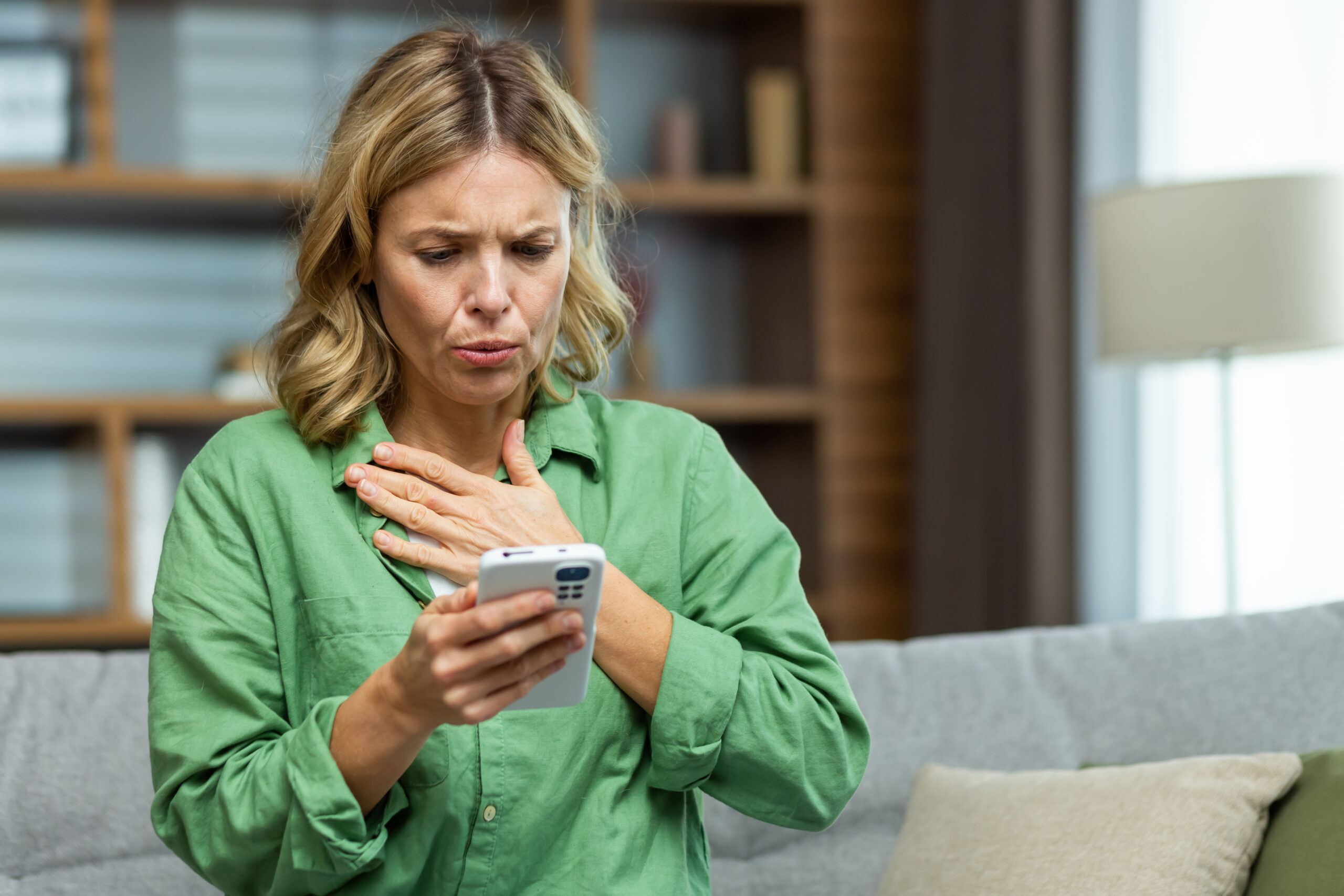 Lonely mature woman alone close up holding phone upset and depressed reading bad news on smartphone online in living room close up.